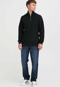 Schwarzer gerippter Strick-Zipfelpulli mit hohem Kragen. Kombiniert mit dunkelblauen Jeans und weißen Sneakers mit schwarzen Streifen.