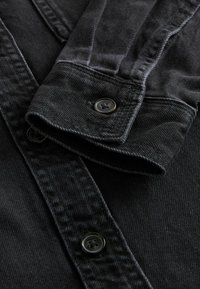 Donkerdenim shirt met een opgerolde mouw die een zwarte knoop, knoopsgat en zichtbare stiksels op het stofoppervlak toont.