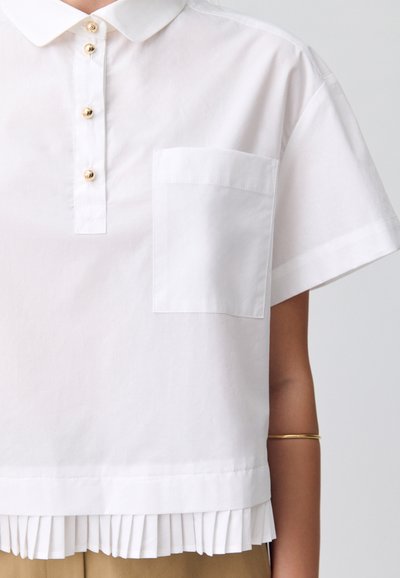 Camisa blanca de manga corta con cuello, botones dorados y un bolsillo en el pecho. Presenta un dobladillo plisado para mayor textura.