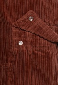 Roestkleurige corduroy stof met zichtbare drukknoopsluitingen, met de nadruk op een open flapdetail.