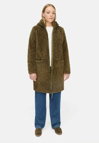 Bruine, fuzzy-gebreide jas met capuchon, ritsluiting en voorzakken. Gecombineerd met losse blauwe jeans en bruine laarzen.