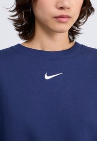 Navy modra majica z dolgimi rokavi, izdelana iz mehke tkanine, z belim Nike swoosh logotipom na prsih in rebrastim ovratnikom.