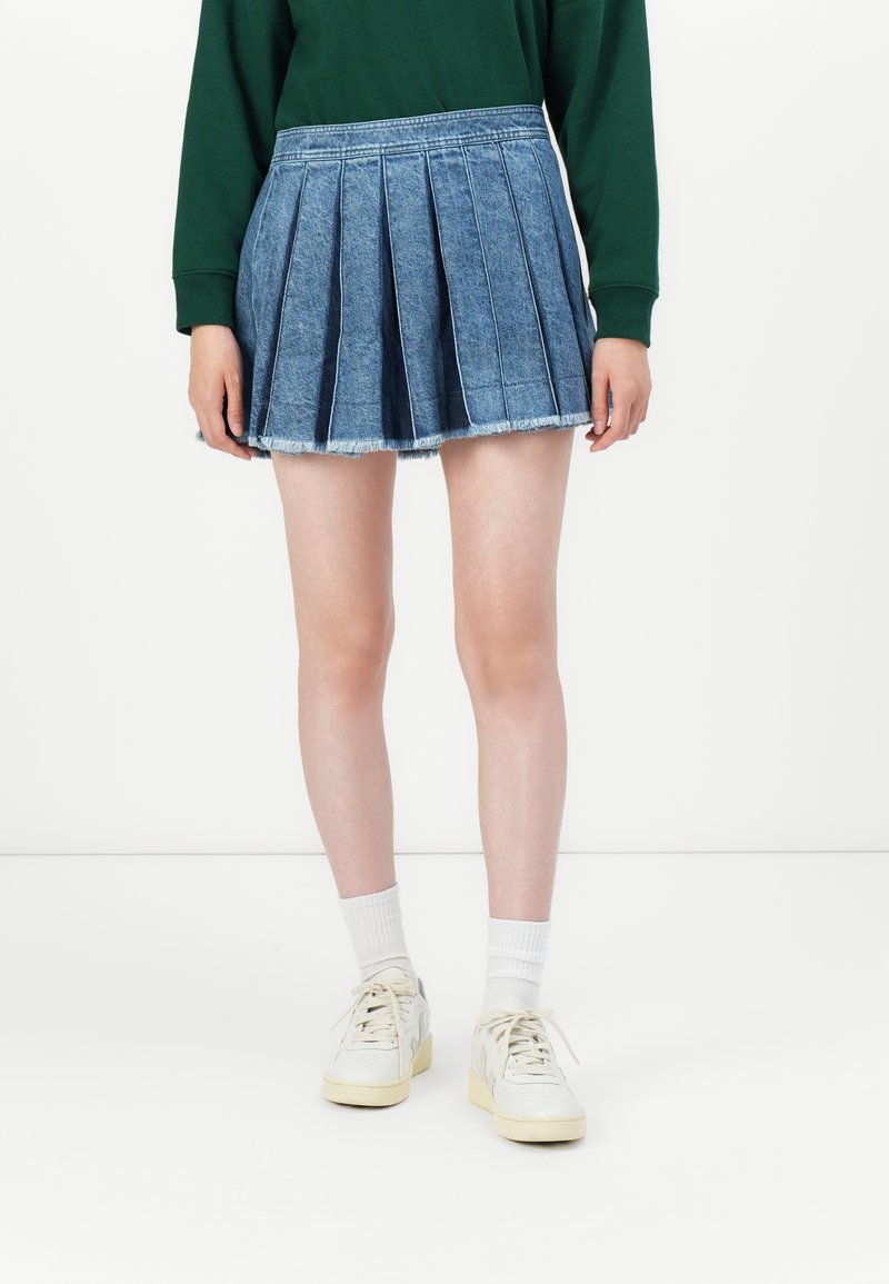 Una persona indossa una gonna di jeans blu plissettata, scarpe da ginnastica bianche e calzini bianchi, in piedi contro uno sfondo bianco semplice.