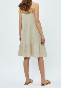 Beige bomullsklänning med justerbara spaghettiremmar, lagerdesign och volangkant, med en texturerad yta. Bärs med platta sandaler.