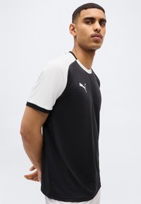 Junger Mann steht mit den Händen hinter dem Rücken, trägt ein schwarz-weißes Puma-Sportshirt und blickt leicht nach oben und zur Seite.