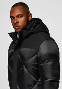 Giubbotto imbottito nero con design trapuntato, dotato di cappuccio, chiusura con zip e patch logo sulla manica. Texture liscia e lucida.