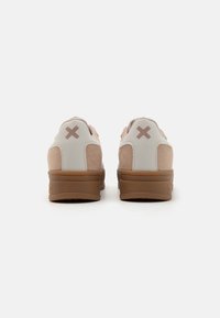 Zapatillas de ante rosa con un talón de cuero blanco, suela de goma marrón y un distintivo logo marrón en forma de "X" en la parte trasera.