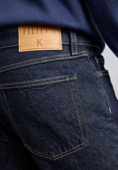 Gros plan sur la poche arrière d'un jean en denim bleu foncé avec une étiquette de marque en cuir marron cousue au-dessus de la poche, sur une personne portant un pull marin.