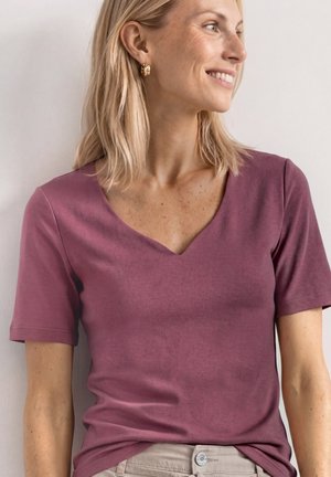 Kobieta o blond włosach, nosząca złote kolczyki koła i krótką bluzkę z głębokim dekoltem w kolorze mauve, uśmiechająca się i patrząca na bok.
