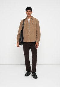 Bruine corduroy button-up shirt met twee borstzakken, gecombineerd met een donkerbruine broek en zwarte schoenen. Model draagt een zwarte schoudertas.