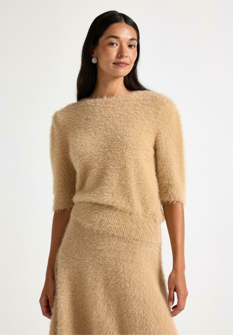 Beige, mjuk tröja med korta ärmar, med en vid halsringning och en strukturerad yta. Kombineras med en matchande kjol som framhävs.