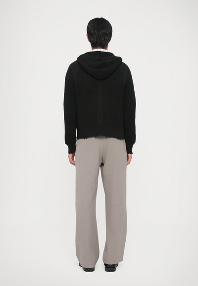 Svart stickad hoodies med en strukturerad yta och huva, kombinerad med ljusgrå byxor. Sömmen är fransad, vilket ger en avslappnad detalj.