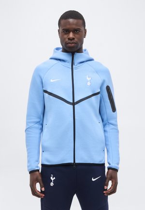 Mann trägt hellblauen Hoodie mit Reißverschluss und Tottenham Hotspur- sowie Nike-Logos, kombiniert mit dunkelblauen Nike-Hosen mit passenden Logos.