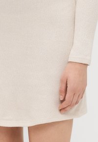 Robe en tricot beige clair avec une texture côtelée. Elle présente des manches longues et une coupe décontractée, mettant en valeur une main reposant près de l'ourlet.