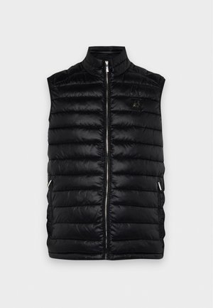 Un homme porte un bob noir, un gilet matelassé, un haut à manches longues, un pantalon foncé et des mocassins sur un fond clair uni.