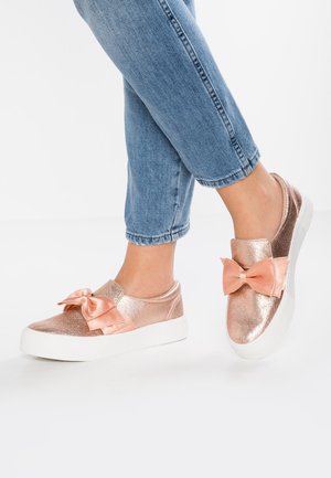 Růžově zlaté metalické slip-on boty s saténovou mašlí, bílou podrážkou a hladkým povrchem. Nosené s ohrnutými modrými džínovými kalhotami.