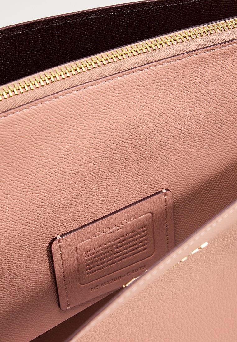 Interiör av en rosa läderhandväska från Coach som visar en guldfärgad dragkedja och en insydd läderetikett med präglad text och serienummer.