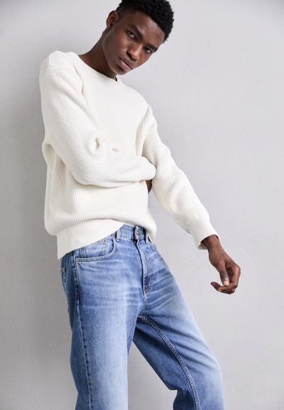 Krém színű bordázott kötött pulóver, amelyet világoskék, egyenes szárú farmernadrággal párosítottak. A pulóver hosszú ujjú és kerek nyakkivágású.