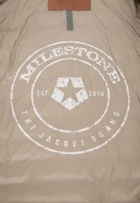 Beige Jackeninnenfutter mit einem runden, verblassten weißen Logo und dem Text "MILESTONE EST. 1994 THE JACKET BRAND" sowie einem braunen Lederlabel.