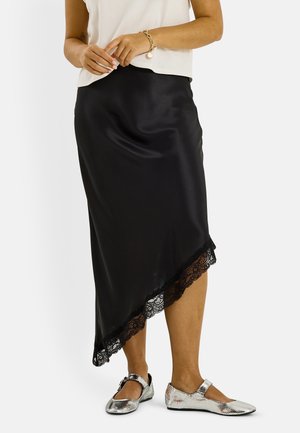 Mujer con falda de raso negra asimétrica con dobladillo de encaje, camiseta blanca sin mangas, pulsera dorada y zapatos planos plateados estilo Mary Jane.