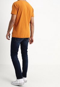Orange bomull t-shirt med korta ärmar, kombinerad med mörkblå jeans. Personen har vita sneakers, sett från ryggen.