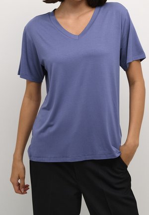 Vrouw draagt een blauw T-shirt met korte mouwen en V-hals, met zwarte broek, één hand in de zak, tegen een effen lichte achtergrond.