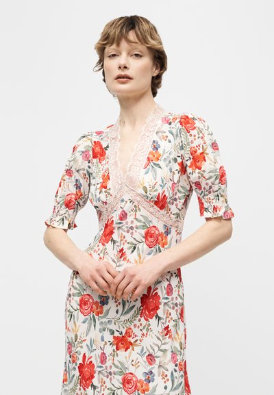 Γυναίκα με λευκό φόρεμα με λουλούδια σε κόκκινο, ροζ και πορτοκαλί χρώμα, με δαντελένια διακόσμηση και φουσκωτά μανίκια, που στέκεται μπροστά σε απλό φόντο.