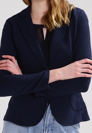Femme portant un blazer bleu marine sur un haut noir, bras croisés, associée à un jean bleu clair, sur un fond uni.
