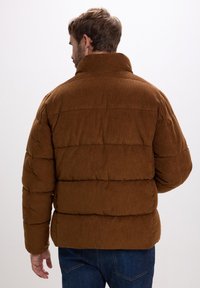 Giacca puffer in velluto a coste marrone con colletto alto, caratterizzata da sezioni trapuntate e una vestibilità rilassata. Indossata con jeans in denim blu.