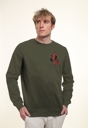 Junger Mann trägt ein olivgrünes Sweatshirt mit Ski-Grafik und dem Schriftzug „SKI STYLE“, steht vor hellem, einfarbigem Hintergrund.