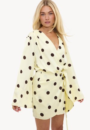 Femme blonde portant une robe portefeuille jaune pâle à pois noirs et manches longues larges, debout devant un fond blanc.