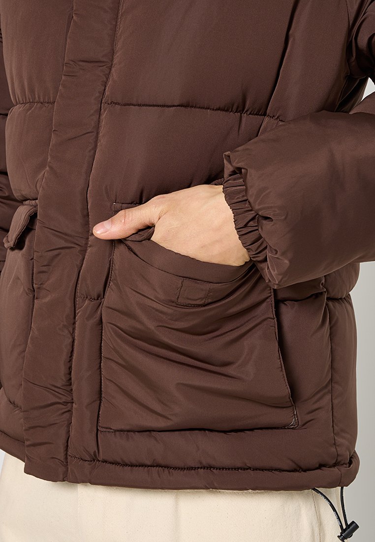 Braune Pufferjacke mit einer weichen, glänzenden Textur. Verfügt über elastische Bündchen, Seitentaschen und einen leichten Glanz, der eine voluminöse Form verleiht.