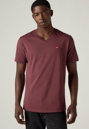 Νέος άνδρας που φοράει απλό μπορντό T-shirt με V-neck και κοντά μανίκια με μικρό κόκκινο λογότυπο Levi's στο αριστερό στήθος, συνδυασμένο με μαύρο παντελόνι.