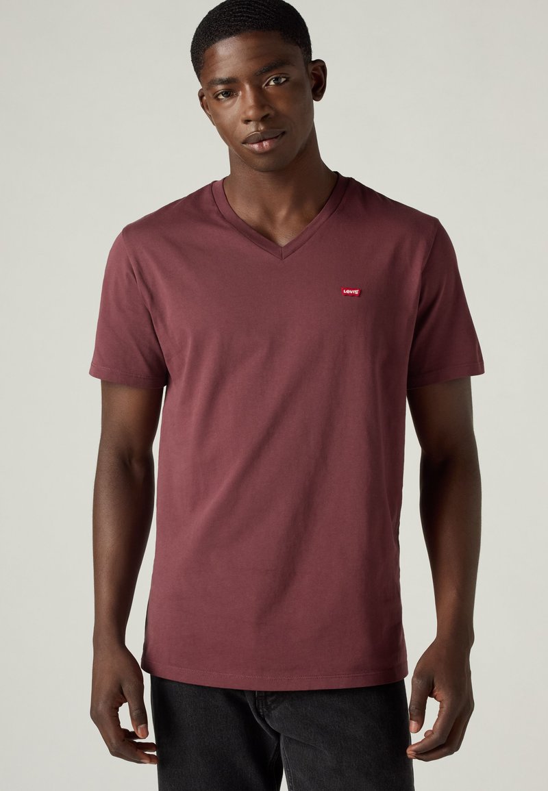 Νέος άνδρας που φοράει απλό μπορντό T-shirt με V-neck και κοντά μανίκια με μικρό κόκκινο λογότυπο Levi's στο αριστερό στήθος, συνδυασμένο με μαύρο παντελόνι.