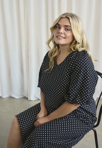 Robe noire à pois fabriquée en tissu léger, dotée d'un décolleté en V et de manches ballons. Le modèle est assis sur une chaise devant un fond blanc.