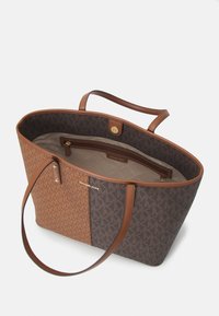 Sac fourre-tout marron présentant un mélange de motifs monogrammes et de textures unies, un intérieur beige et des accessoires dorés. Dispose de deux longues poignées en cuir.