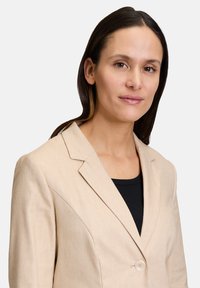 Beżowa lniana marynarka z jednolitym zapięciem na guzik, klapami z wycięciem oraz lekką teksturą, zestawiona z czarną dopasowaną bluzką pod spodem.