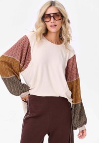 Długosleeve top w kolorze kremowym z wzorzystymi, kolorowymi rękawami w burgundzie, musztardzie i szarości. Posiada strukturalny splot; noszony z brązowymi spodniami.