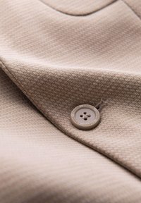 Gros plan d’un tissu beige texturé avec un bouton cousu, embossé du mot "OMBRE" autour de son bord.
