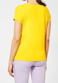 Žluté tričko s krátkým rukávem a kulatým výstřihem, hladká textura a kontrastní šití. Spárováno s světle fialovými kalhotami.