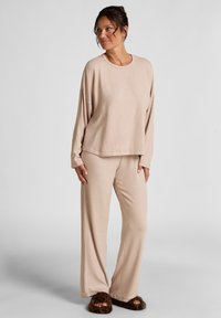 Licht beige loungewear set van zacht materiaal; bevat een ontspannen pasvorm top met lange mouwen en wijde broekspijpen, gecombineerd met bruine pluizige slippers.