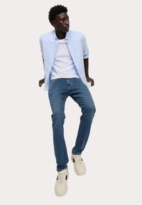 Camicia button-up azzurro chiaro sopra una maglietta bianca, abbinata a jeans blu e sneakers crema con accenti verde scuro.