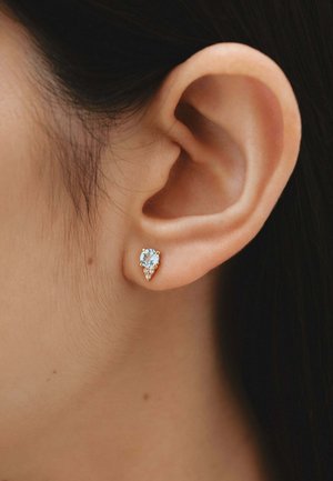 Pendiente de stud en tono dorado que presenta una gema azul en forma de gota, adornada con pequeñas piedras transparentes a su alrededor.