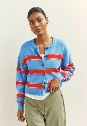 Frau, die eine blaue Strickjacke mit breiten roten und weißen Streifen, beige Hosen und goldene Creolen trägt, steht vor einem schlichten hellen Hintergrund.