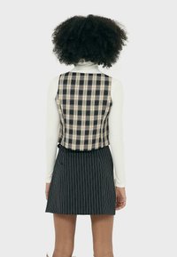 Vue arrière d'une personne portant un gilet en plaid noir et crème sur un haut à manches longues blanc, associé à une jupe noire à rayures fines.