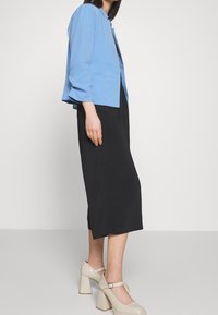 Světle modrá cropped bunda s dlouhými rukávy, nošená přes černou midi sukni, doplněná o nahé boty na blokovém podpatku. Hladká látka a propracovaný střih.