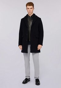 Cappotto in lana blu navy con rever a lancia, chiusura con bottoni e due tasche anteriori, indossato sopra una giacca in pelle nera, abbinato a pantaloni grigi e scarpe nere.