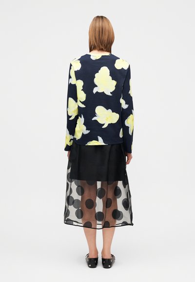 Μπλούζα με μακριά μανίκια σε μπλε ναυτικό με κίτρινα σχέδια, συνδυασμένη με διάφανο μαύρο φούστα με πουά. Μαύρα παπούτσια ολοκληρώνουν το σύνολο.