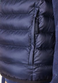 Tmavě modrá prošívaná vesta ve stylu puffer s kapsou na zip na boku a visačkou s logem Lacoste, vyrobená z hladkého, lesklého materiálu.