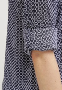 Chemise à manches longues avec un motif géométrique bleu marine et blanc, manche retroussée, montrant l'avant-bras à la peau claire.